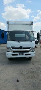 Front view of a 2019 Hino 155 box truck showing windshield, headlights, grille, and cab; white exterior with cargo box above.
