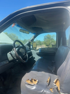 Interior view of a 2000 Ford F550 cab and chassis showing damaged front bench seat with torn upholstery, steering wheel, dashboard, and window in the background.