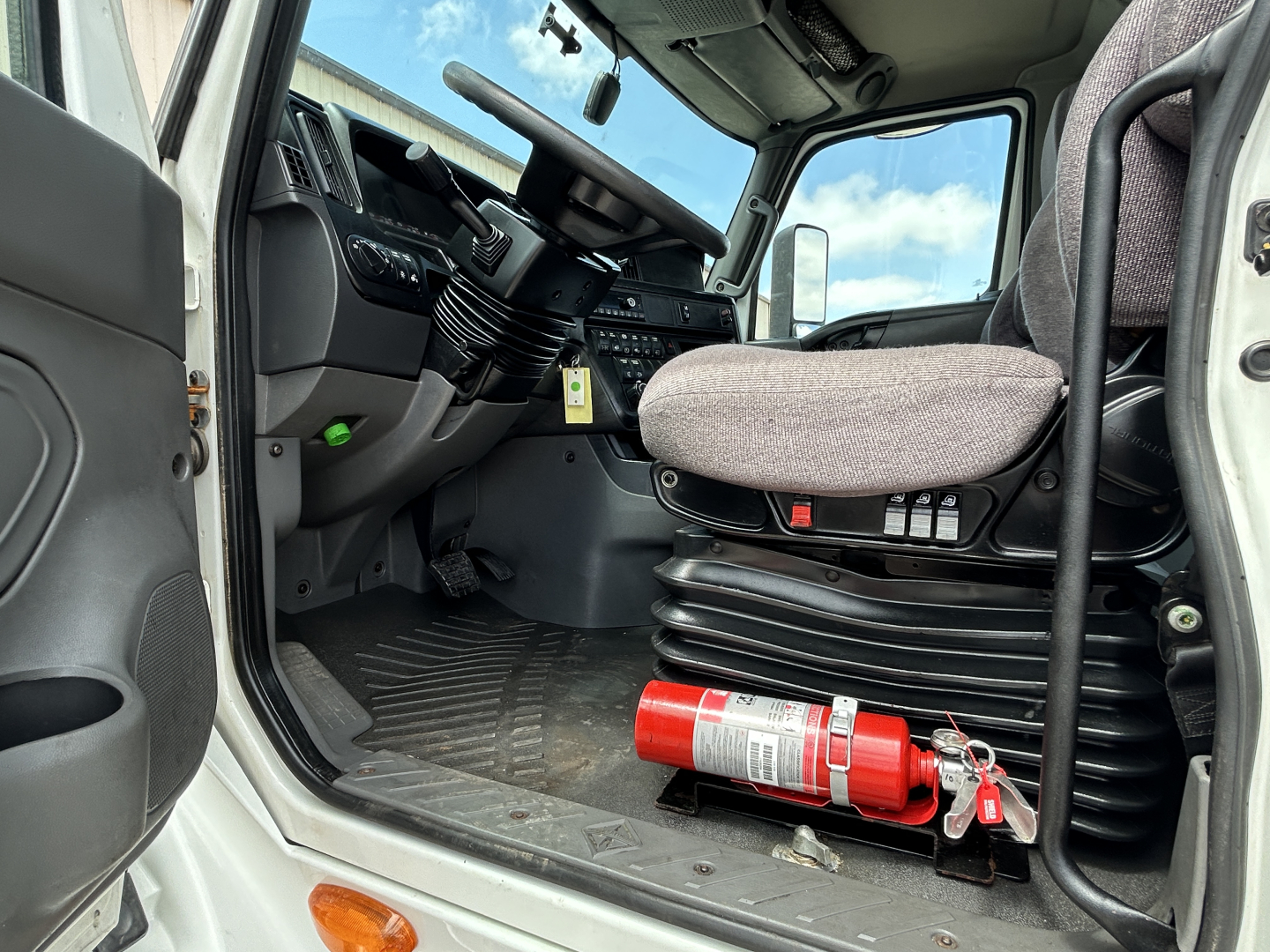 Truck cab interior showing driver's seat, steering wheel, and floor with a red fire extinguisher mounted near the door sill.