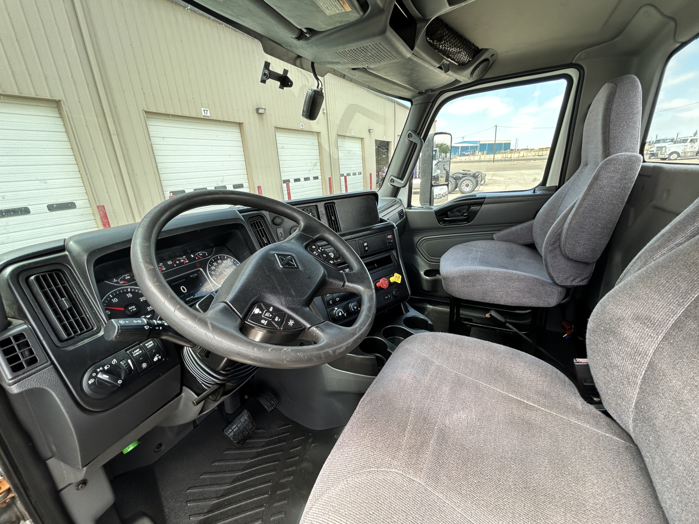 Interior of 2021 International LT625 truck cab with steering wheel, dashboard, and grey cloth seats. View shows driver's seat, passenger seat, and center console.