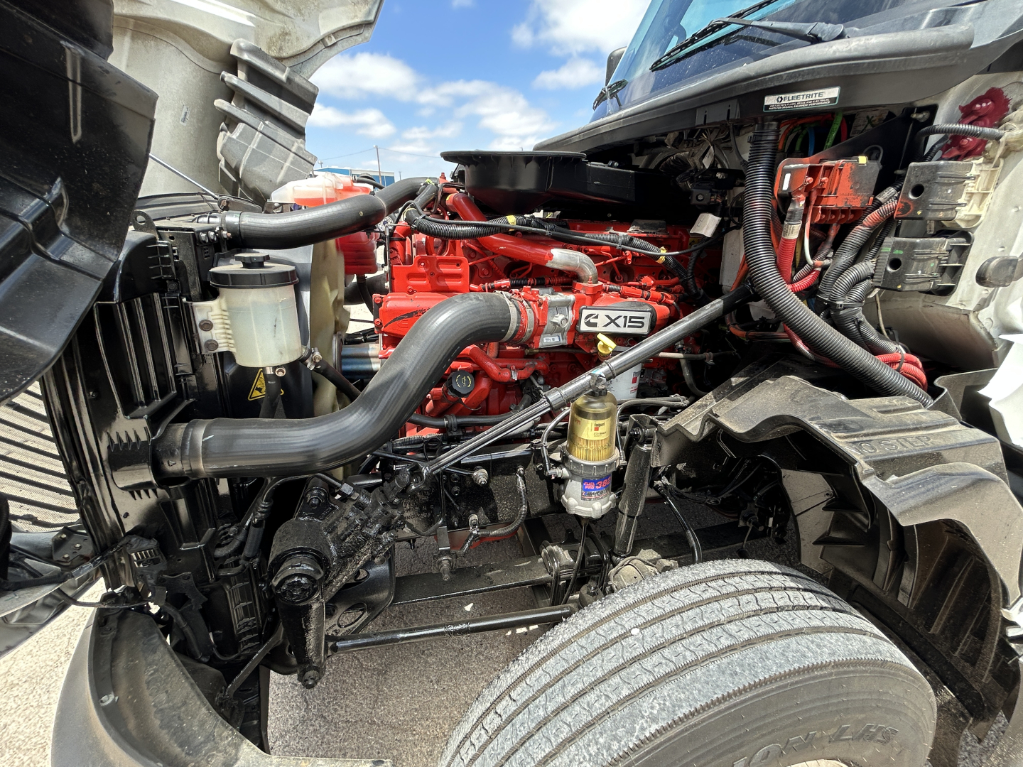 Truck engine bay showing Cummins X15 engine with red block, hoses, radiator, and various components.