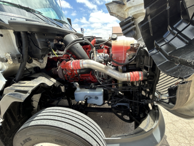 Truck engine bay of a 2021 International LT625, showing red engine components, silver turbo/pipework, coolant reservoir, front suspension, and exposed tires.