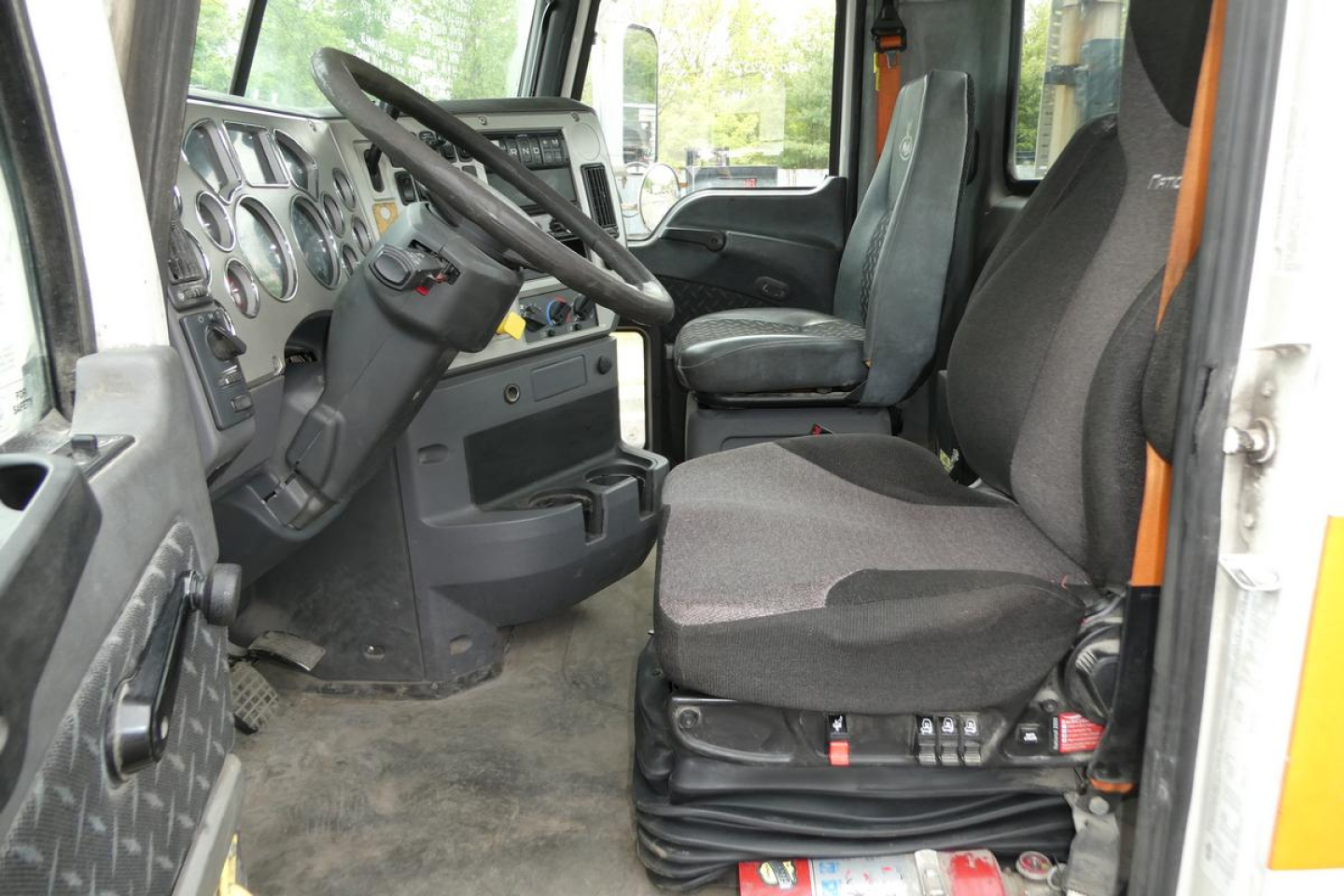 Interior of a 2018 Mack CXU613T cab showing steering wheel, dashboard gauges, driver seat, and passenger seat with seat controls visible.