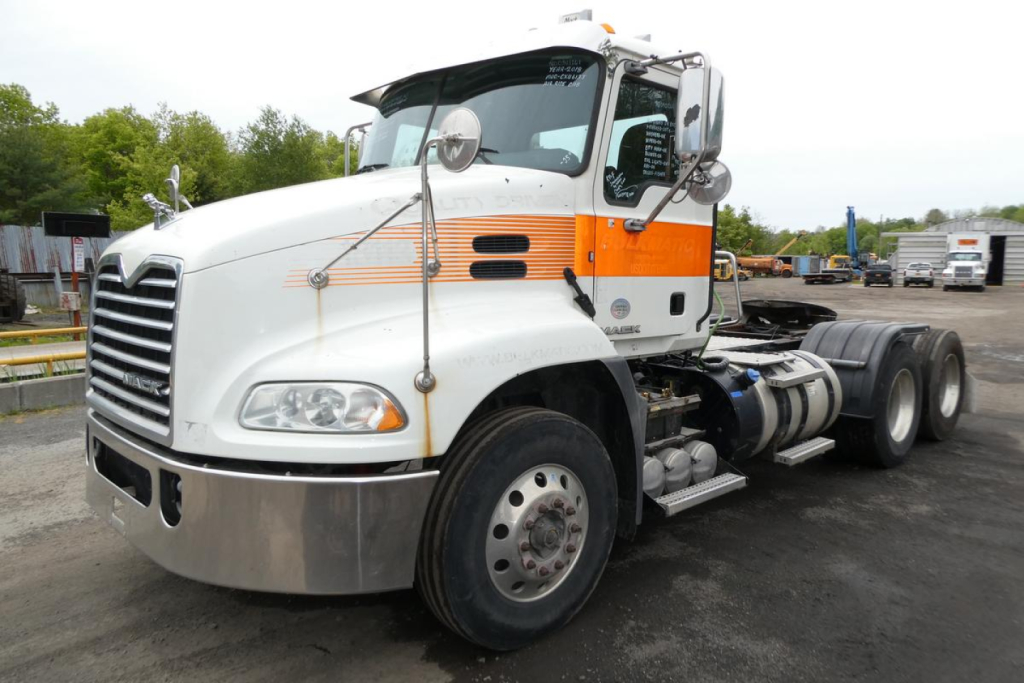 Truck cab and chassis of a white 2018 Mack CXU613T with orange stripe, showing front grille, headlights, and dual rear wheels.