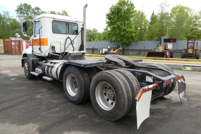 Truck chassis of a 2018 Mack CXU613T daycab tractor from rear left angle; visible components include dual rear axles with large tires, white cab with orange reflective stripes, exhaust stack, and trailer hitch area.