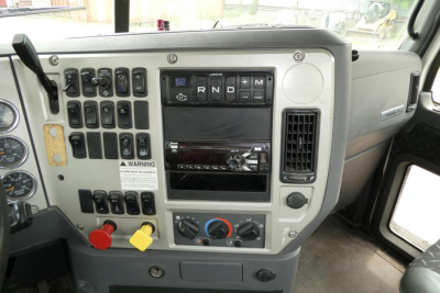 Dashboard of Mack CXU613T interior showing gauge cluster and controls, gear selector with R N D P, multimedia screen, climate controls, and auxiliary switches.