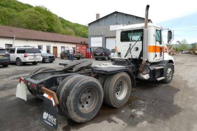 Truck chassis and cab of a used Mack CXU613T tractor with dual rear axles, white cab with orange stripe, parked in a lot.