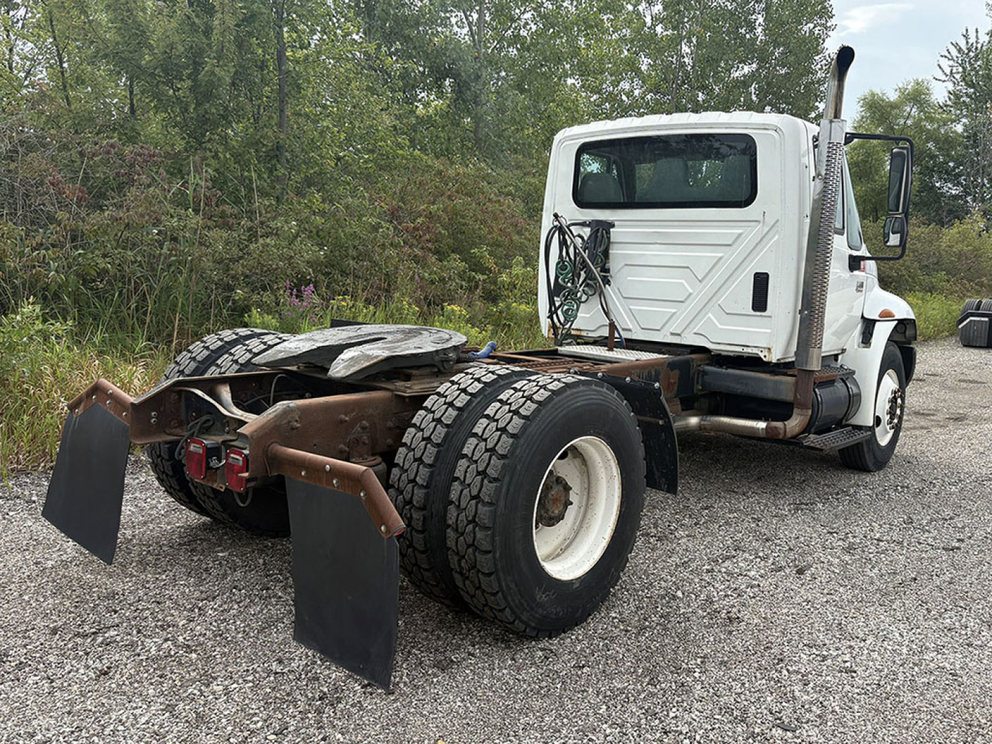 Truck chassis with rear dual wheels and mudflaps, white cab, bare frame, no trailer attached, vegetation in background.