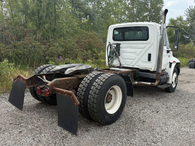 Truck chassis with rear dual wheels and mudflaps, white cab, bare frame, no trailer attached, vegetation in background.