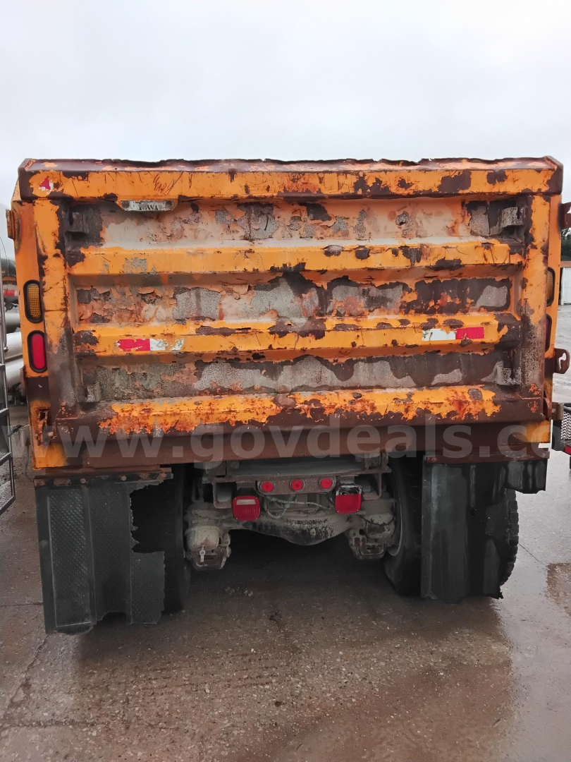 Back of a yellow Mack GU dump truck with a rusted bed, visible red taillights, and rear axles; weathered body panels and muddy tires.