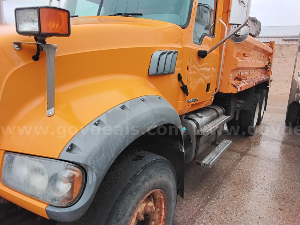 Orange Mack GU Granite dump truck front cab and wheel area showing side steps and fender; visible text on side panel and headlight assembly.