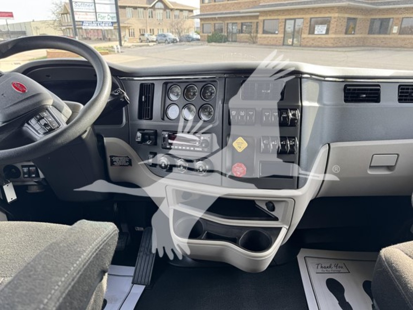 Peterbilt 579 dashboard with steering wheel and instrument cluster; visible labels include Peterbilt badge on the steering wheel, various gauges, switches, and cup holders; showroom/lot background visible.
