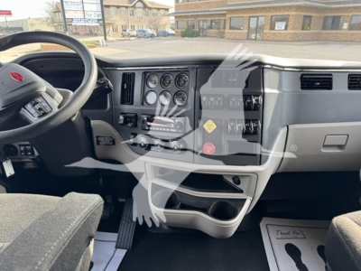 Peterbilt 579 dashboard with steering wheel and instrument cluster; visible labels include Peterbilt badge on the steering wheel, various gauges, switches, and cup holders; showroom/lot background visible.