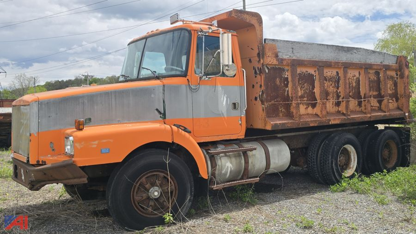 Dump truck side view showing orange cab with gray grille and a rusted dump bed, three-axle arrangement, fuel tanks under the cab, and worn wheels.