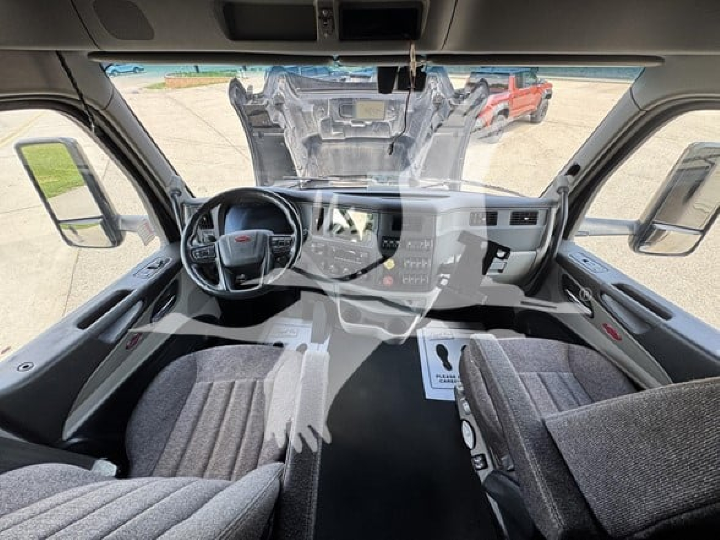 Truck cab interior of a 2023 Peterbilt 579, driver seat, steering wheel, dashboard, center console, seats, and windshield view of engine bay area reflected as overlay