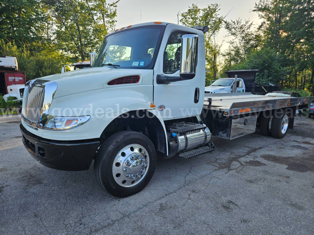 2019 International MV607 tow truck with white cab and flatbed. Headlights, grille, large side mirrors, fuel tanks under cab, dual rear wheels. VIN/stock text not on image itself.
