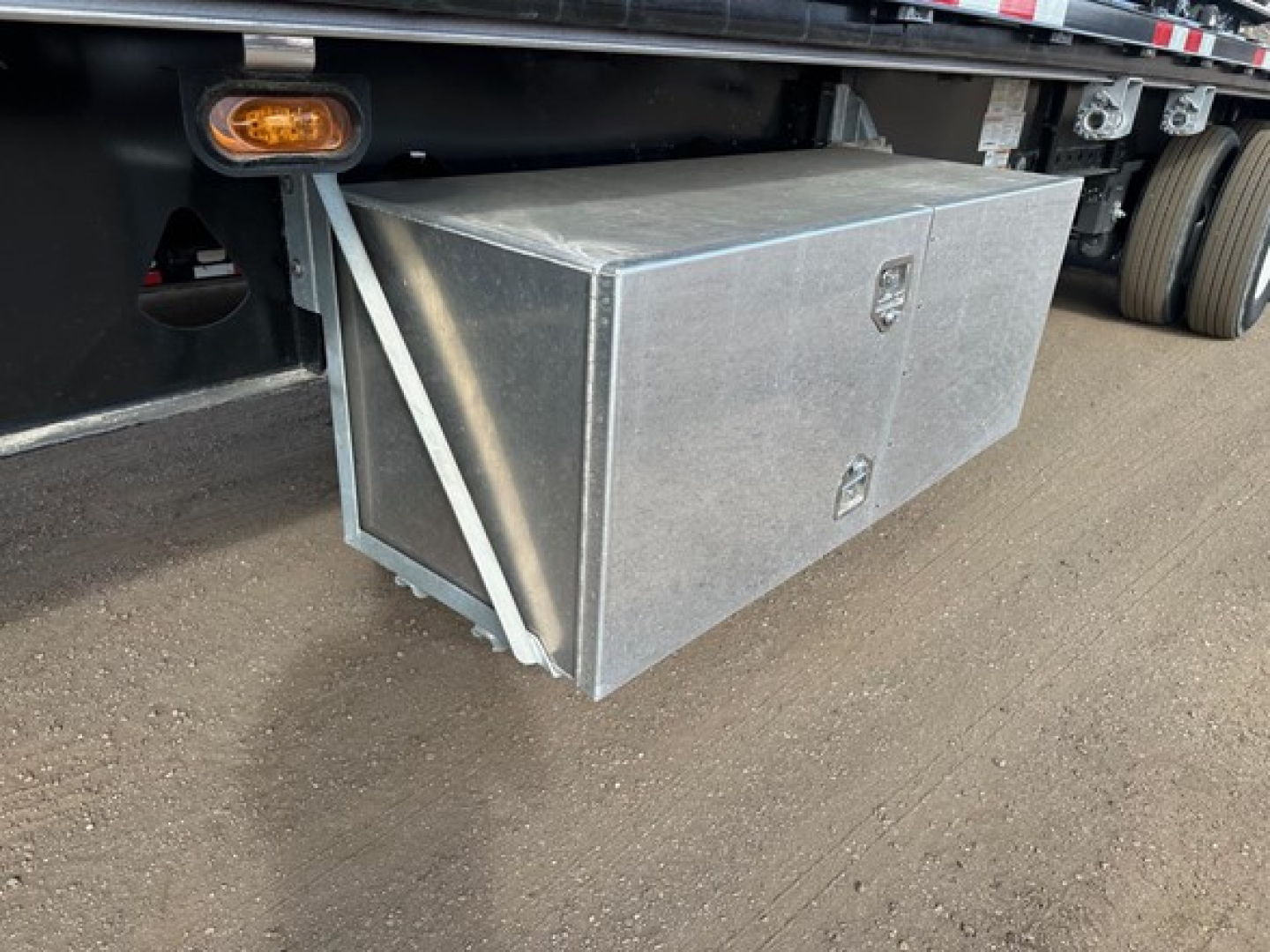 Flatbed trailer toolbox mounted under side with two-latch aluminum storage box and rearmost wheel area visible.
