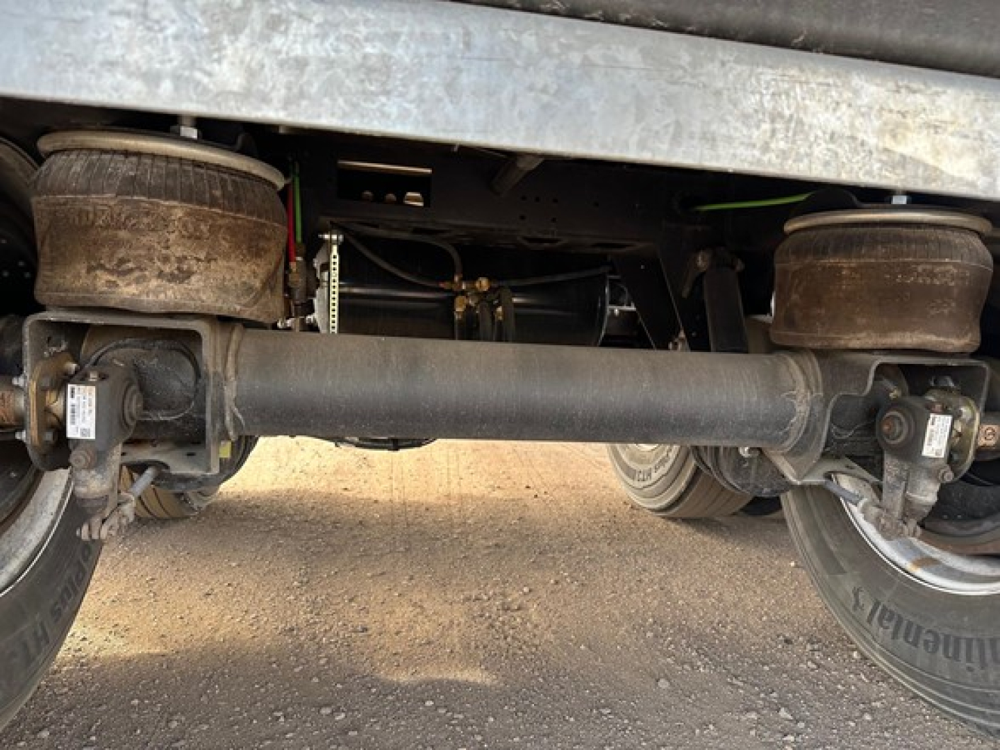 Underneath a 2026 Manac 48' Flatbed trailer showing the rear suspension and drive axle, with leaf springs and air springs visible; no legible text readable.