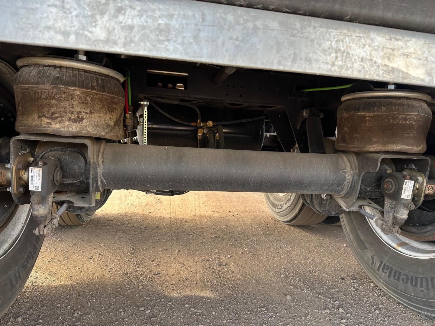 Trailer underside showing axle housings, suspension, and drive shaft of a flatbed trailer. Visible tires and leaf springs with dirt on drums and hubs.