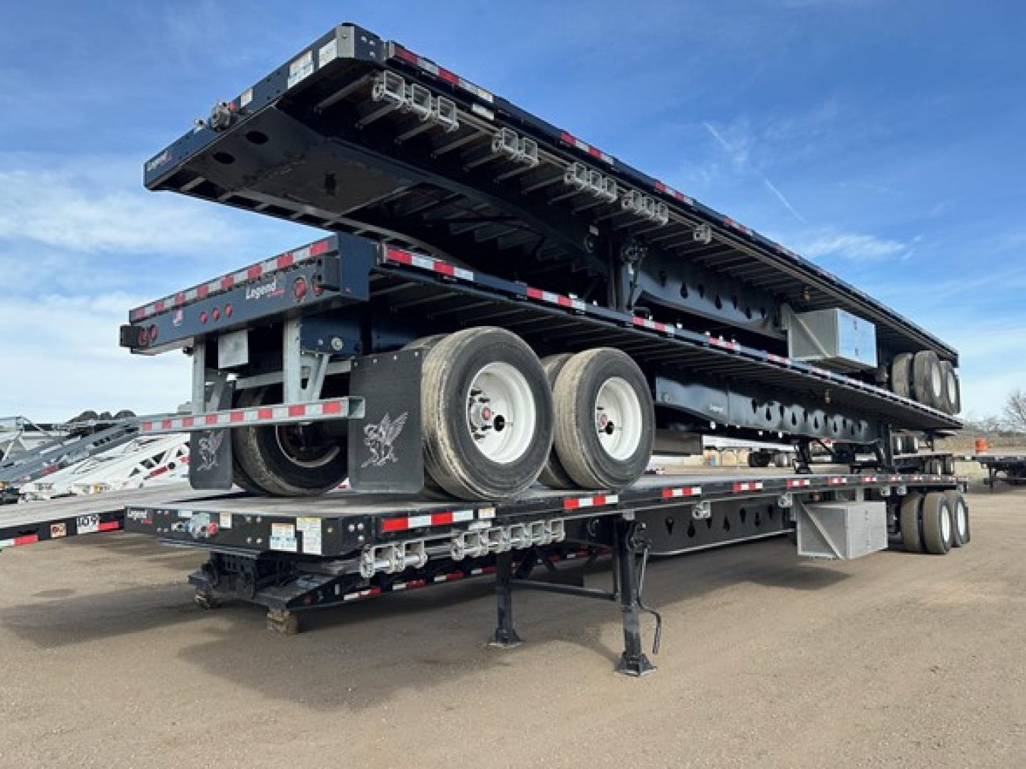 Manac steel 48' flatbed trailer with air ride and air pin slide, triple-axle configuration, piled tandem wheels, stacked loading ramps, side rails, and hydraulic components visible in the open trailer bay.