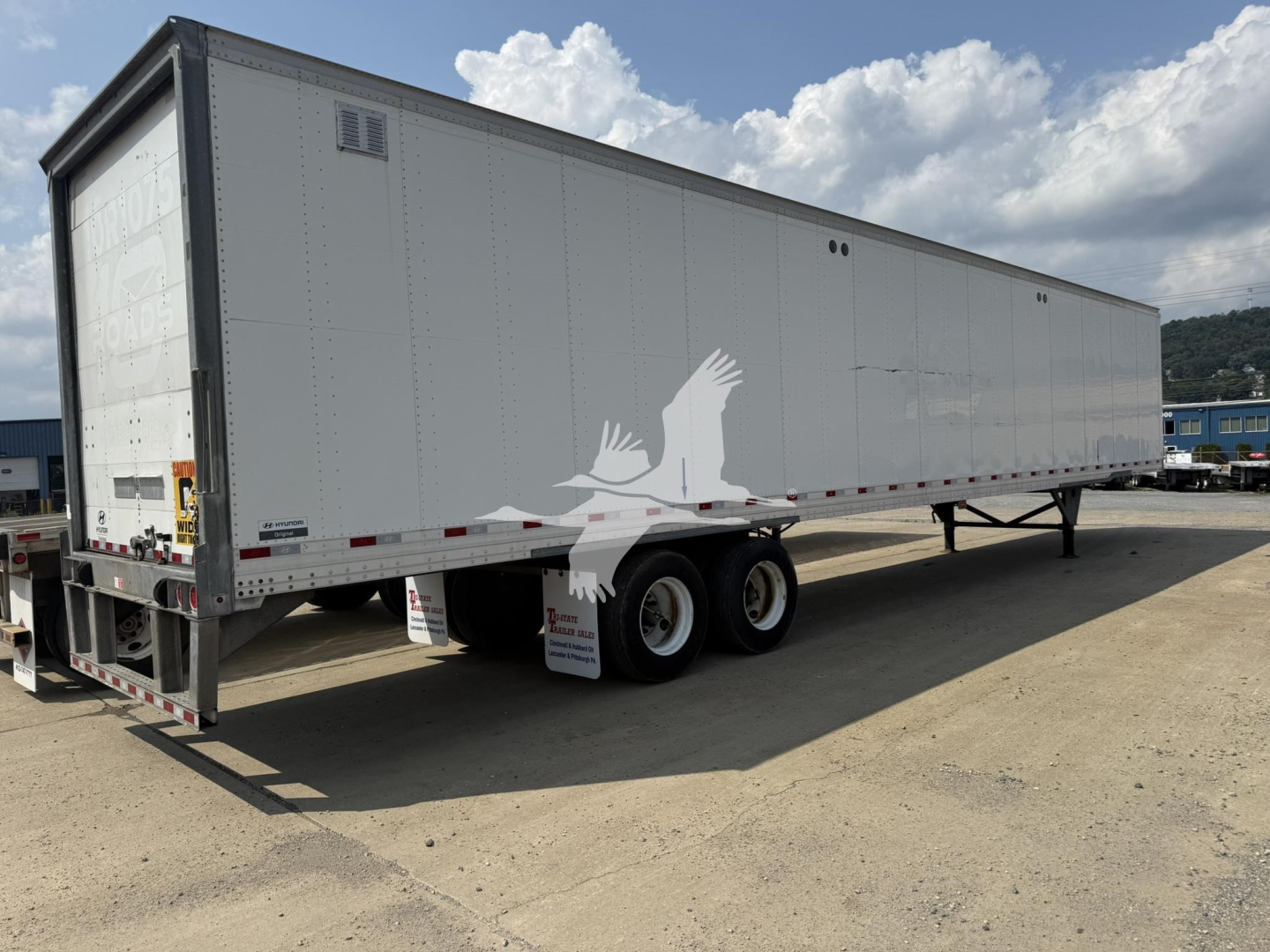 Trailer side view showing a long rectangular white roll door van trailer with two rear axles and black frame. Visible text on rear left door area and small stickers, no license plate.