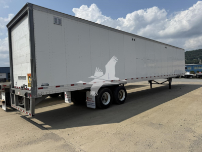 Trailer side view showing a long rectangular white roll door van trailer with two rear axles and black frame. Visible text on rear left door area and small stickers, no license plate.