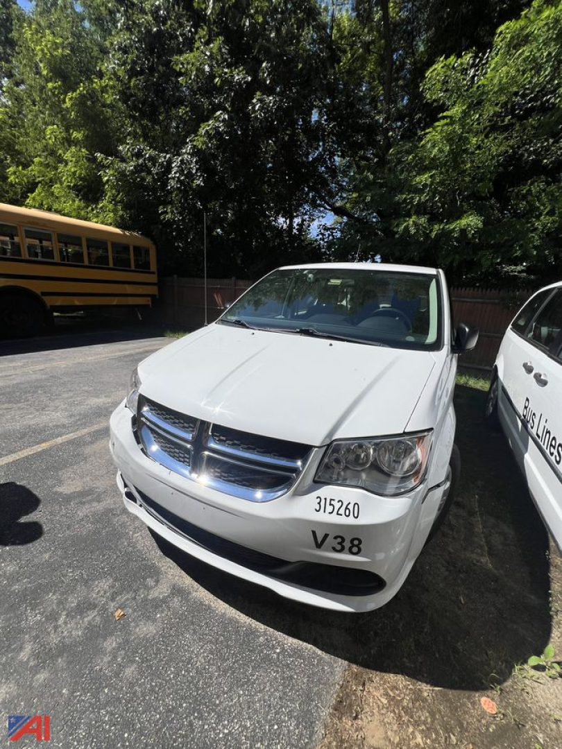 Front view of a white Dodge Grand Caravan minivan with number 315260 and V38 decal on the bumper, parked in a lot near a school bus.