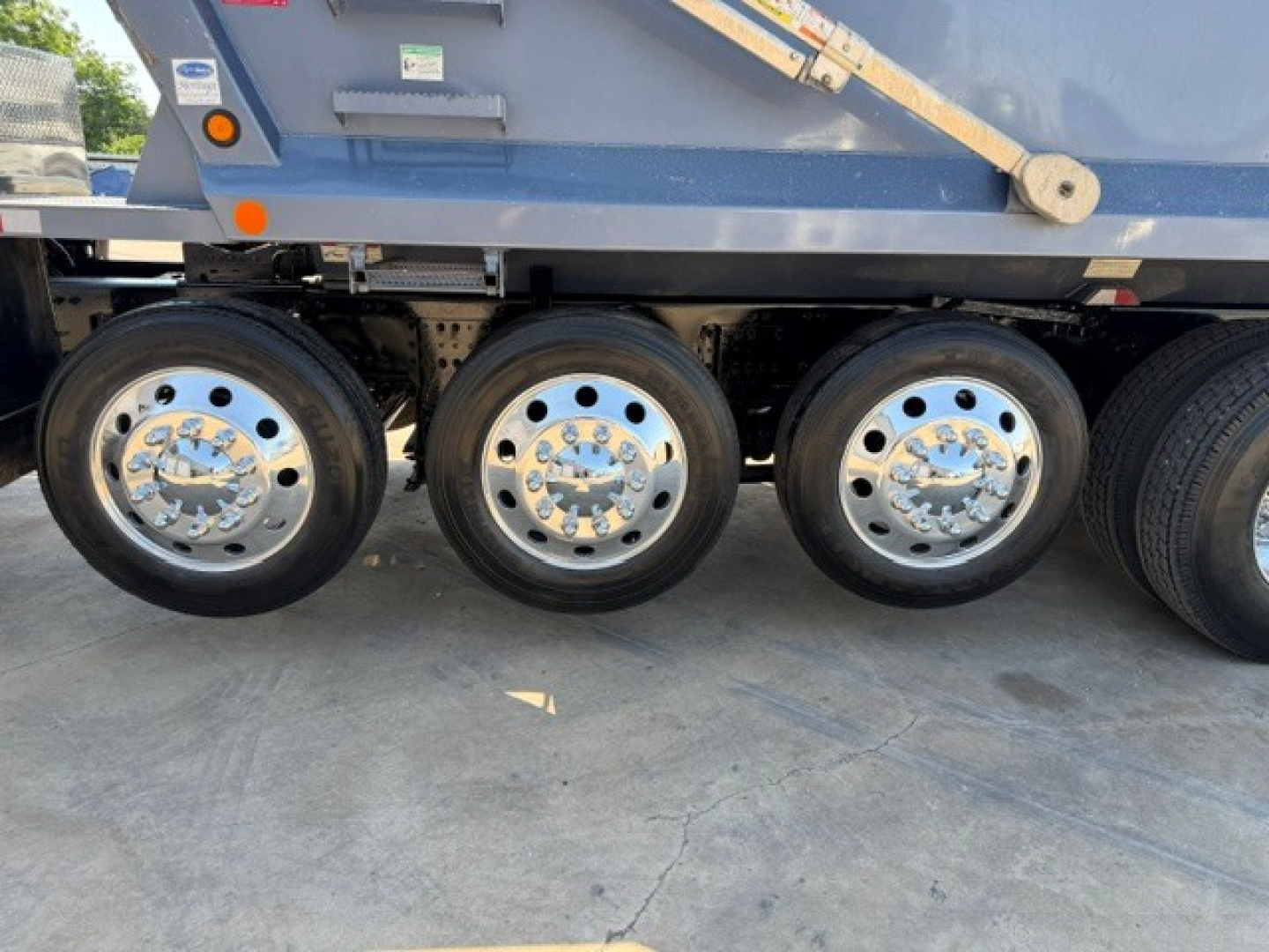 Three rear axles with polished chrome hubcaps on a Volvo VHD truck trailer wheelset, blue side panel above, part of dump truck chassis visible.