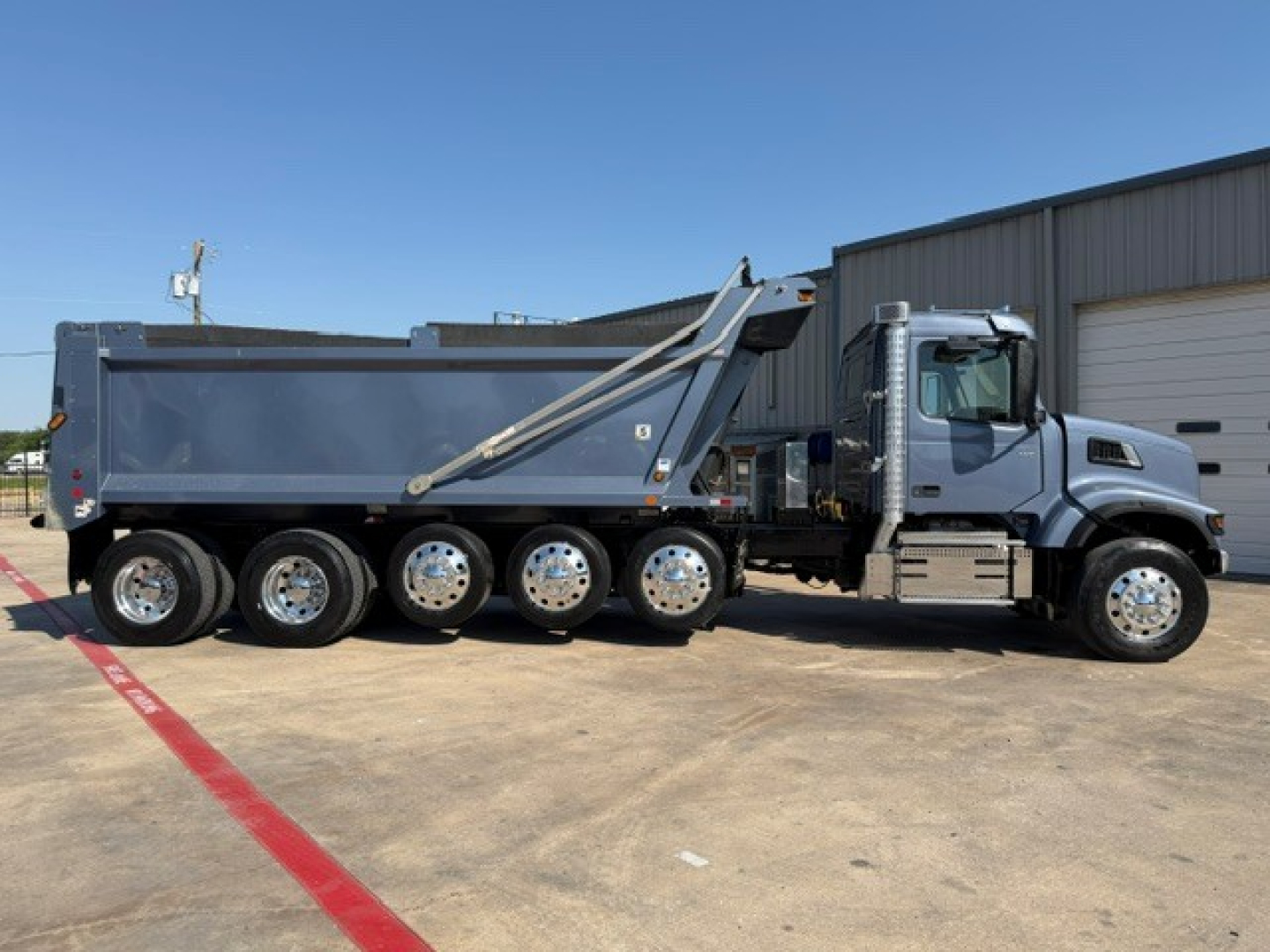 Dump truck side profile showing multi-axle tires under a gray dump bed; Volvo emblem visible on cab, front grille, and hydraulic dump mechanism.