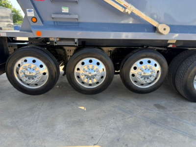 Three rear axles with polished chrome hubcaps on a Volvo VHD truck trailer wheelset, blue side panel above, part of dump truck chassis visible.