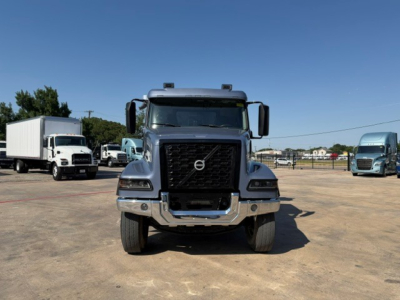 Volvo dump truck front view showing bold grille, large bumper, Volvo emblem, headlights, and side mirrors; clear sky and other trucks in background.