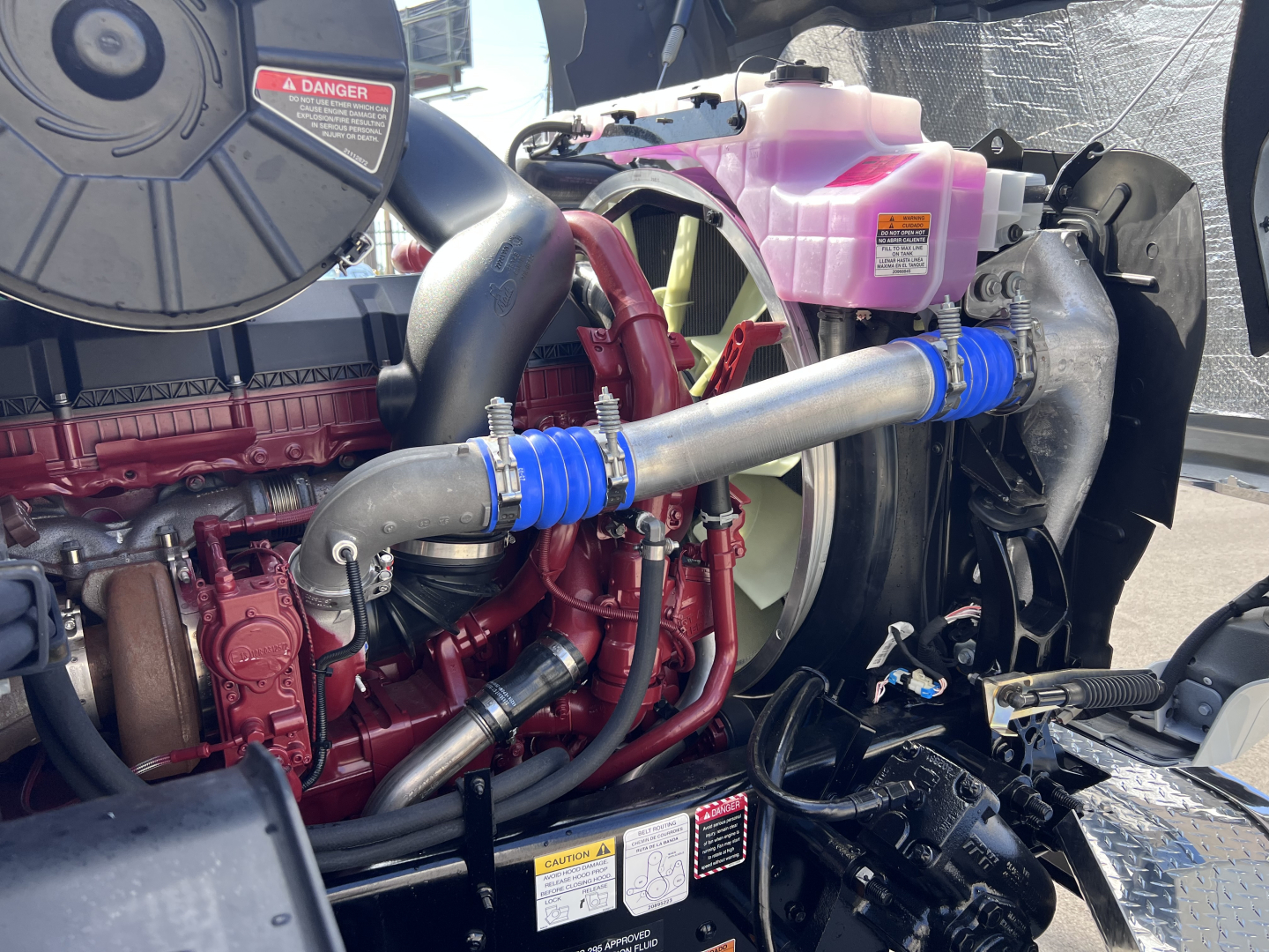 Mack GR64B engine bay with large turbo intercooler piping, pink coolant reservoir, blue silicone hose clamps, and red engine components visible.