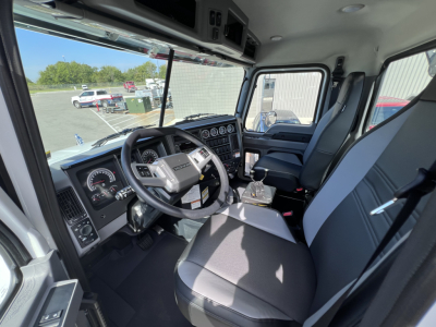 Interior view of a black-and-gray Mack truck cab, driver seat and steering wheel visible, dashboard with gauges and controls, front windshield and side window. 