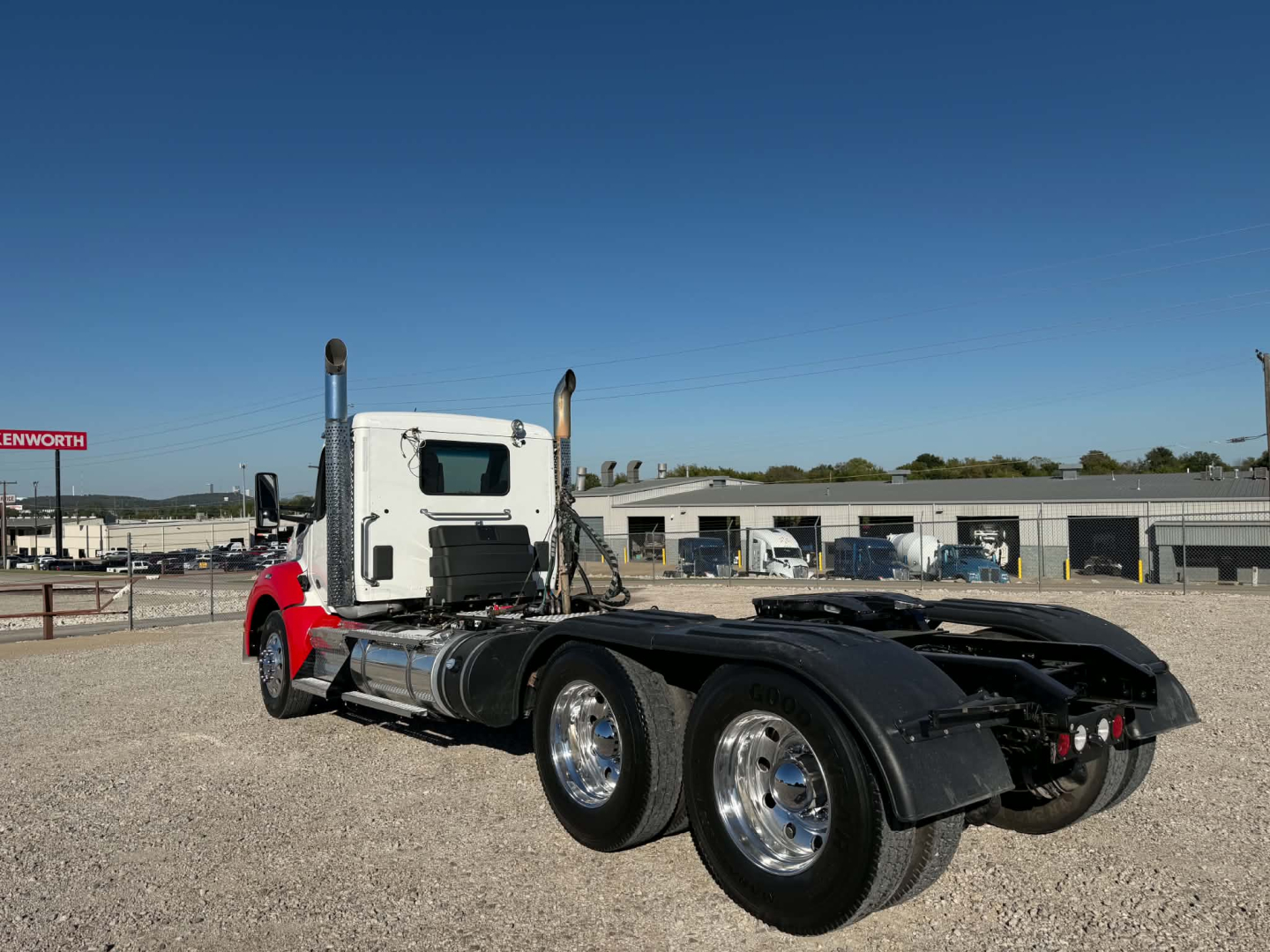 Kenworth T880 conventional daycab truck, side view showing rear dual-axle configuration, chrome wheels, red cab, white sleeper window, gravel lot.