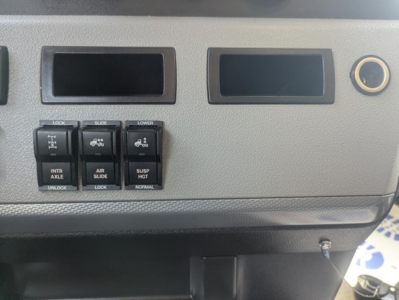 Freightliner dash panel with four small toggle switches labeled INTR AXLE, AIR SLIDE, SUSP HGT, and NORMAL, plus two empty rectangular display slots and a circular cigarette-lighter style outlet.
