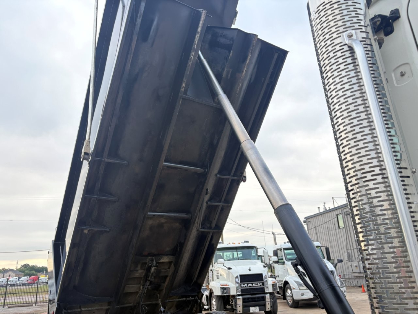 Dump truck bed raised by hydraulic cylinder in a Mack GR64F, with other trucks in background.