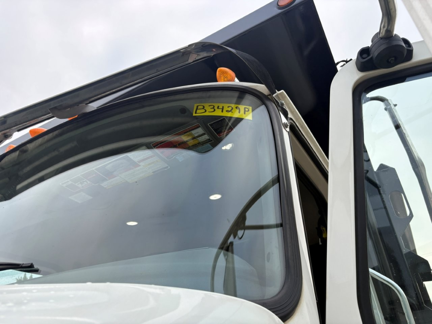 Dump truck cab window with yellow inspection tag reading B3429P and listing sticker area with windshield and door frame.