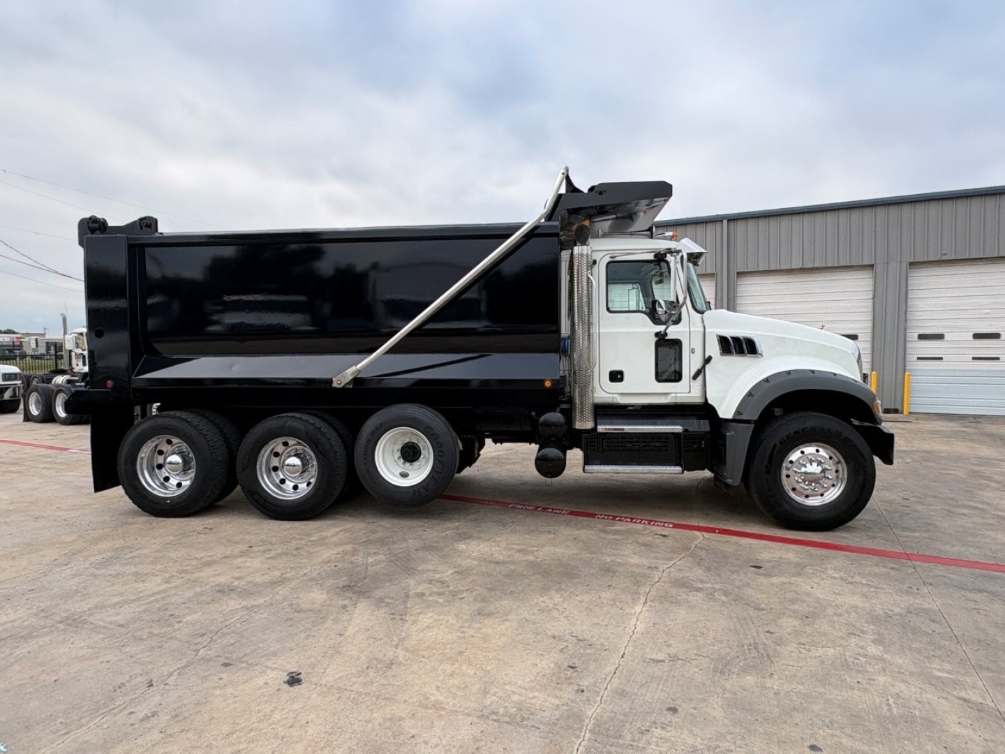 Dump truck side view showing black dump body, white cab, three rear axles, Mack GR64F style with hydraulic dump mechanism and chrome wheels.