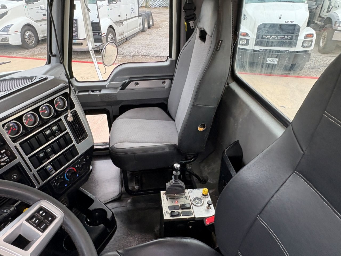 Dump truck cab interior showing driver seat, passenger seat, steering wheel, and gear selector with control panel and switches in a Mack GR64F.