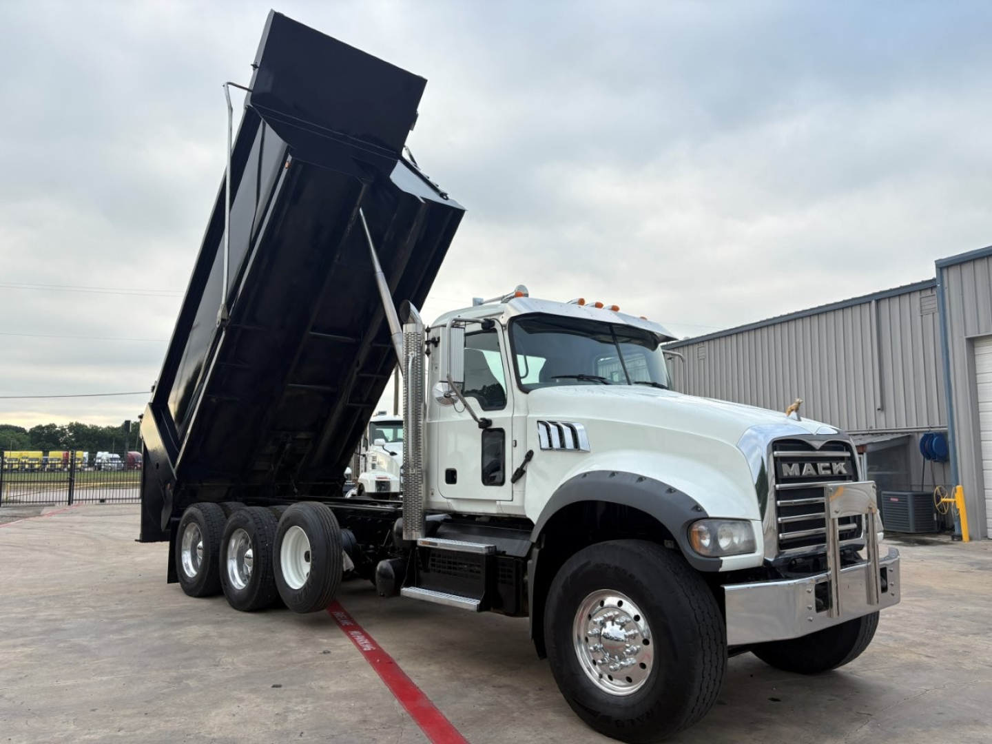 Mack GR64F dump truck front-right side with a raised dumping bed, chrome grille, and dual rear axles in a lot.