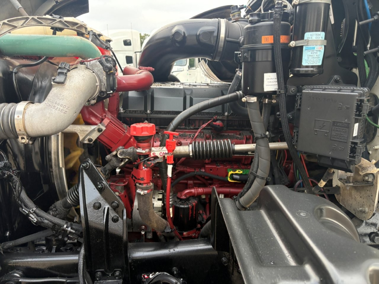 Engine bay of a 2020 Mack GR64F dump truck showing red engine components, hoses, and air intake with surrounding black plastic covers and wiring harnesses.