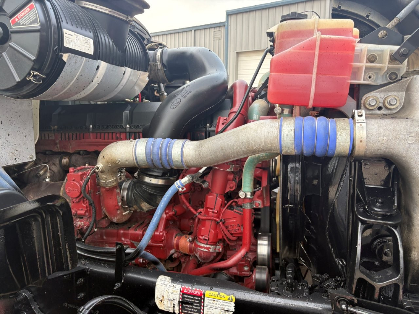 Mack GR64F engine bay showing red diesel engine, blue silicone hoses, black radiator fan, red coolant reservoir, and various metal components.
