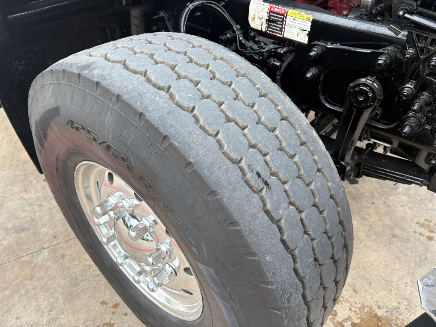 Mack GR64F dump truck front left wheel showing tire tread and chrome wheel, partial engine bay components in background.