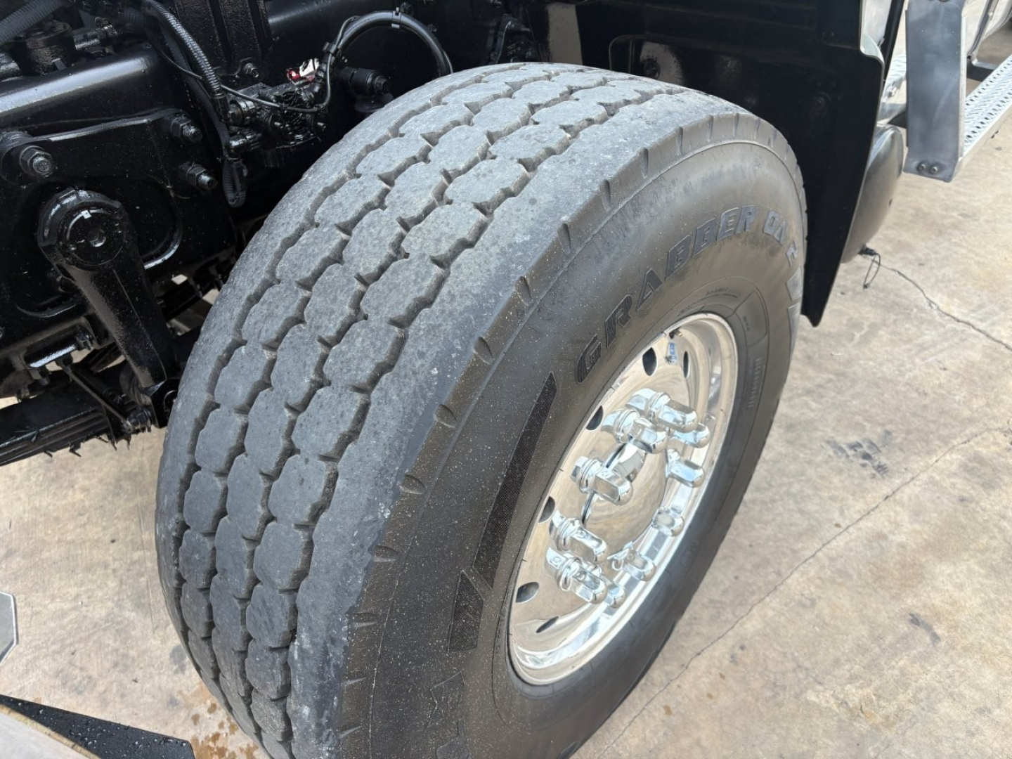 Truck front wheel of a Mack GR64F dump truck showing large treaded tire and polished chrome hub, undercarriage and suspension components visible.