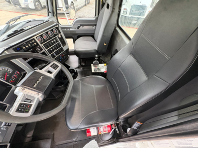 Truck cab interior showing steering wheel with Mack logo, dashboard with gauges, center console, driver seat with seatbelt, gear shifter, and some items on the floor near door.