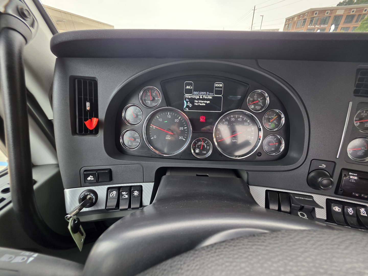 Kenworth T880 dashboard view with multiple gauges and digital display in center; steering wheel at bottom, ignition keys in left; various switches along dash.