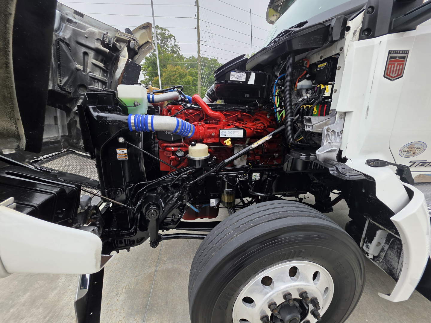 Kenworth T880 engine bay with red engine, blue hoses, and exposed components visible between the cab and front wheel; side of truck shows partial white body panels.