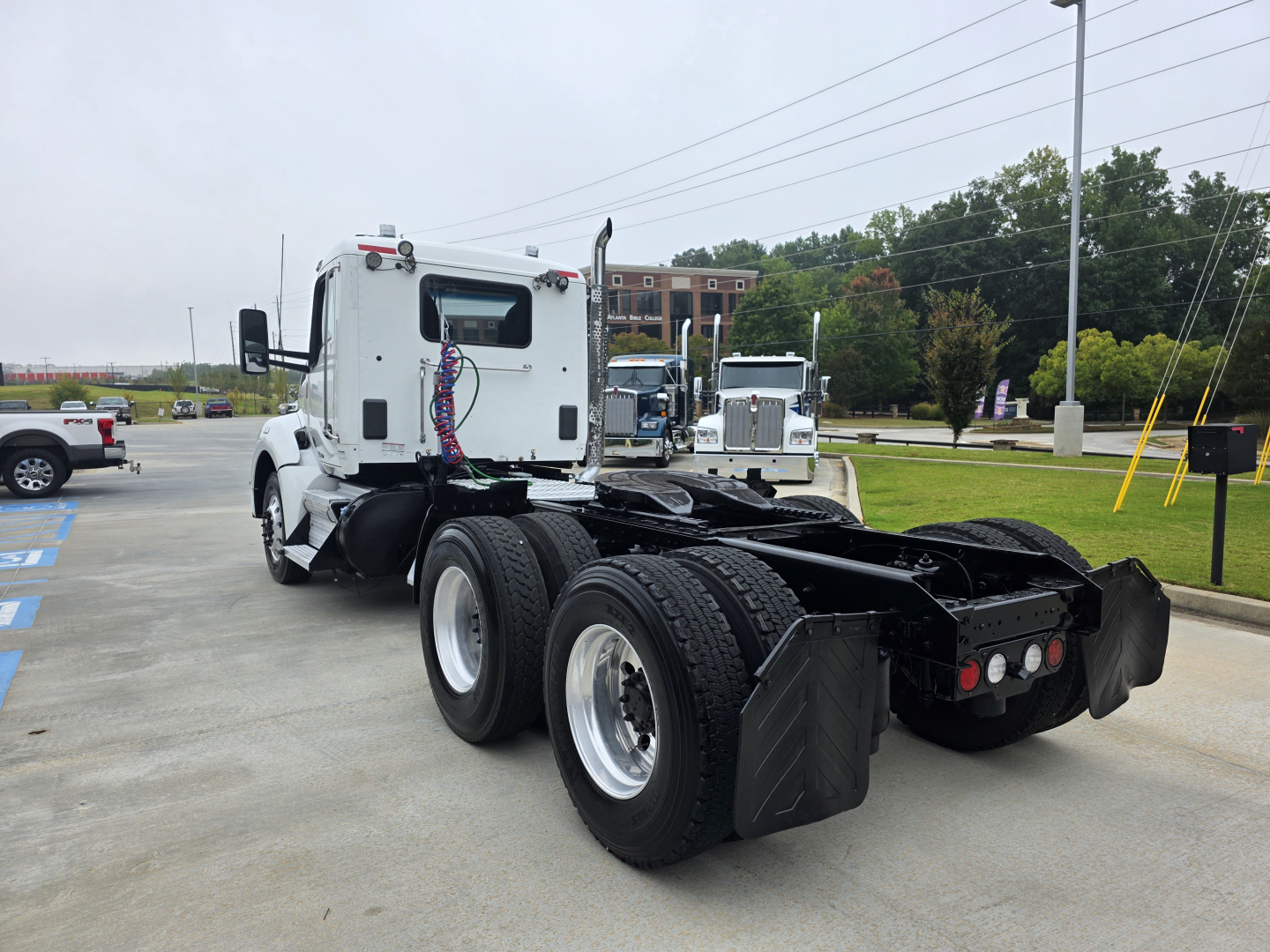 Kenworth T880 tractor in a parking lot, rear view showing dual rear axles and fifth wheel mounting, white cab with hoses and connectors visible.