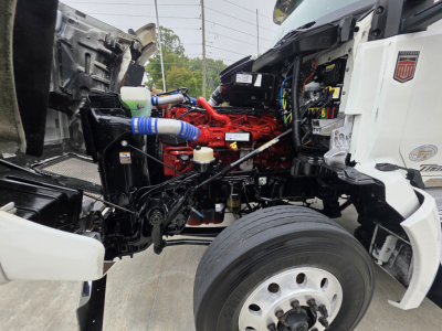 Kenworth T880 engine bay with red engine, blue hoses, and exposed components visible between the cab and front wheel; side of truck shows partial white body panels.