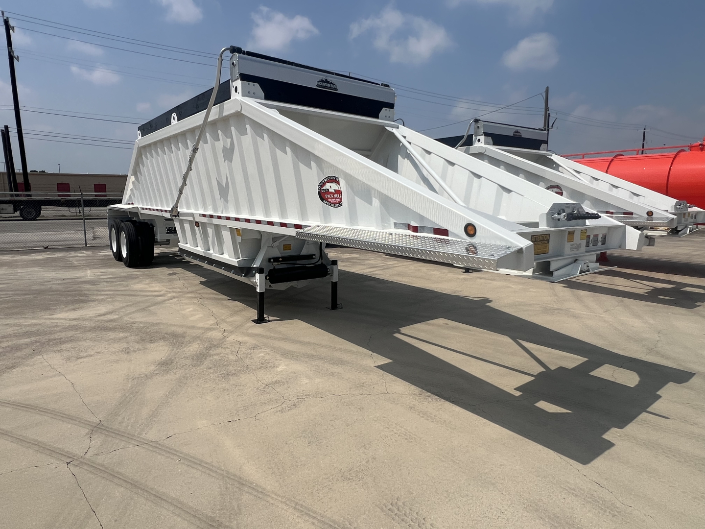 Dump trailer showing white air-ride bottom-dump with dual axles and rear hitch, label on side, and black toolbox on top; side lights visible.